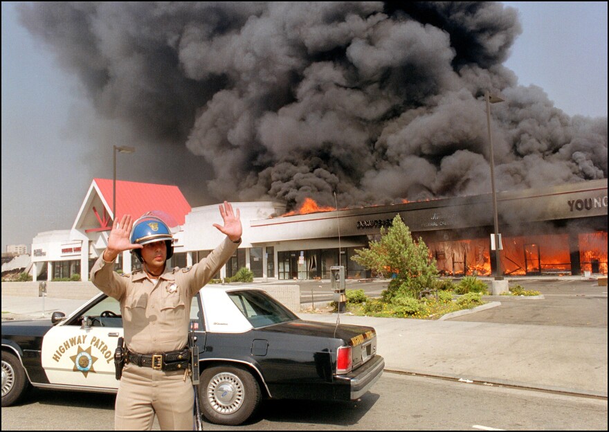 A California Highway patrolman directs raffic around a shopping center engulfed in flames in Los Angeles, 30 April 1992. Riots broke out in Los Angeles, 29 April 1992, after a jury acquitted four police officers accused of beating a black youth, Rodney King, in 1991, hours after the verdict was announced. (Photo credit should read CARLOS SCHIEBECK/AFP/Getty Images)