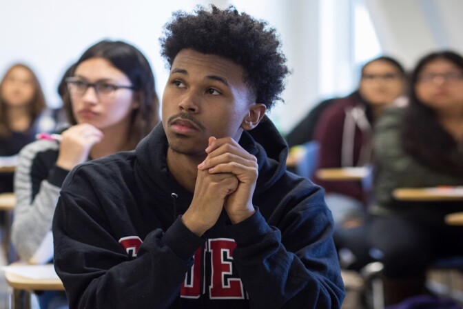 A male college student wearing a dark blue sweatshirt sits at a desk in a classroom, with other students seated at desks in the background.