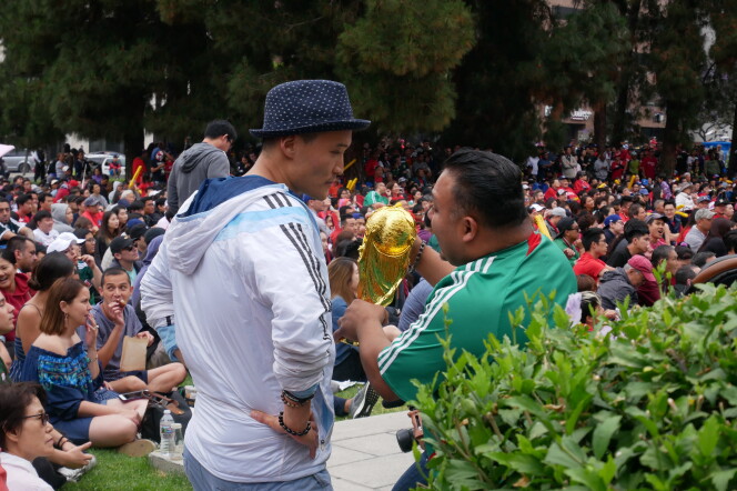 Two men with dark skin stand in front of a crowd talking to each other. One is wearing a light blue jacket and hat with his hands at his side. The other is wearing a green shirt with white stripes on the shoulders while holding a gold trophy. 