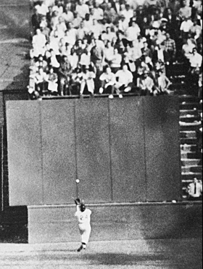A man is running towards a wall and bracing to catch a baseball. A crowd is seen above the man on the field. 