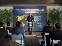 John Kerry speaks at a podium in a dark suit and tie. A map of the world is behind him and people sit in white seats watching him speak. 