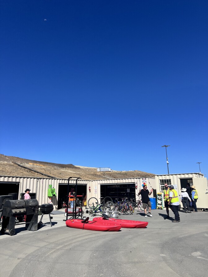 A variety of objects are on display in front of a tan, metal storage-container like structure that's working as a walk-up shop. The objects include two bright red kayaks on the ground, grills, exercise equipment, and bicycles of all shapes and sizes. A man wearing a black shirt and khaki shorts is speaking to two people wearing neon yellow and orange high-visibility vests towards the middle right of the image. 