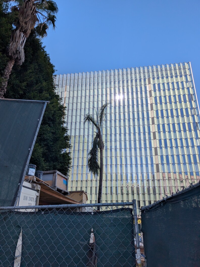 This photo features a modern urban scene with the following elements: A tall building with a glass facade that reflects sunlight, giving it a sleek, contemporary look. In the foreground, there's a construction site enclosed by green mesh fencing, with visible construction materials. Behind the fence is a palm tree.