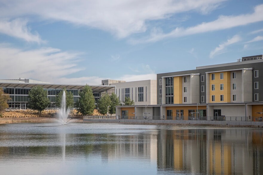 Three- to four-story buildings surround a large fountain on a sunny day. The buildings are painted yellow, dark gray and light gray. They all have rows of windows. A row of five trees is on the far side of the fountain in front of another building.