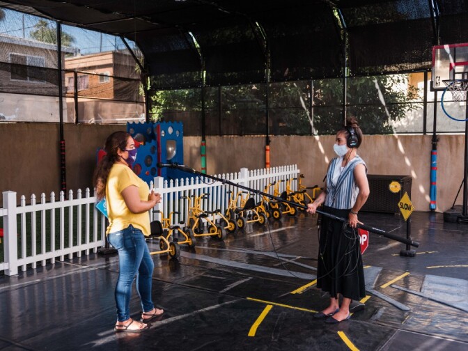 a woman in a yellow shirt and jeans talks into a microphone held on a long boom pole by a woman in a black skirt and striped shirt standing 6 feet away. both of them wear face masks