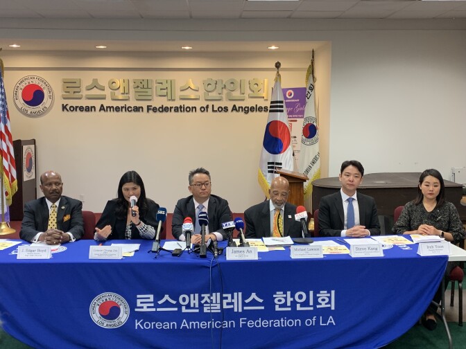 Six Black and Korean American leaders sit at a table together draped in a sign that reads Korean American Federation of L.A. The table is covered with documents and several microphones face the speakers.