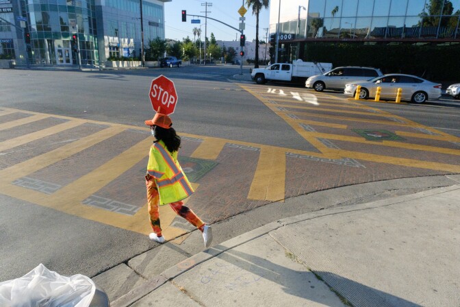 A crossing guard holds a red STOP sign and begins to walk through a busy intersection as drivers queue up in the background.