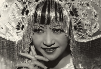 Anna May Wong in her role as Princess Ling Moy. She wears an elaborate headdress that creates a half circle atop her heard, embroidered with beaded filigree. Sparkly fringe falls from the bottom of the headdress framing Wong's face with silver bangs. She smiles looking up from hooded eyes, seemingly micheviously. 