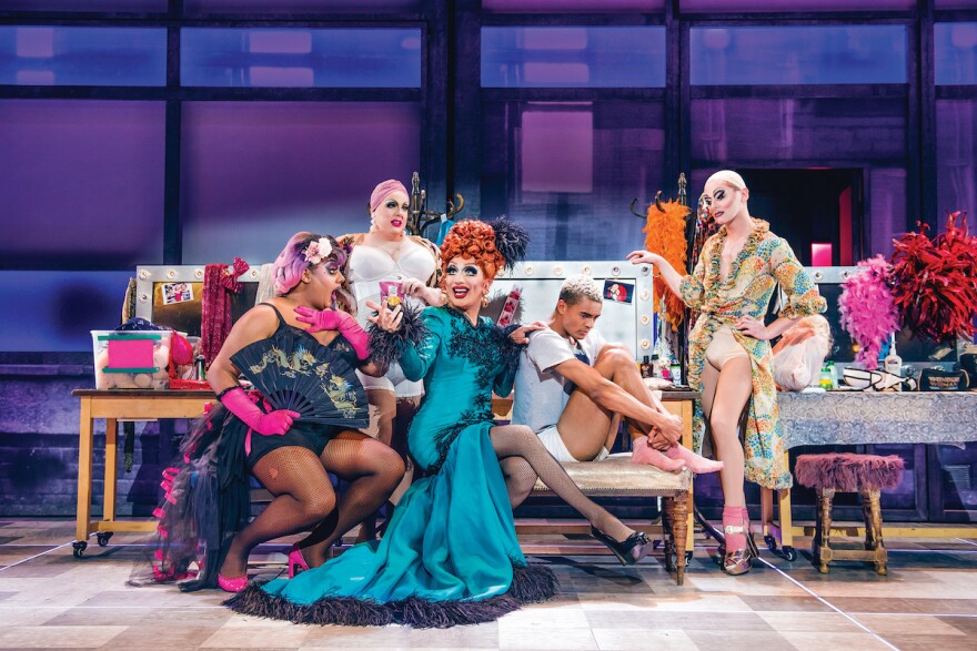 A person with a blue dress and red hair in the center, surrounded by others in various states of undress, on stage with a purple background in what appears to be a simulation of a dressing room.