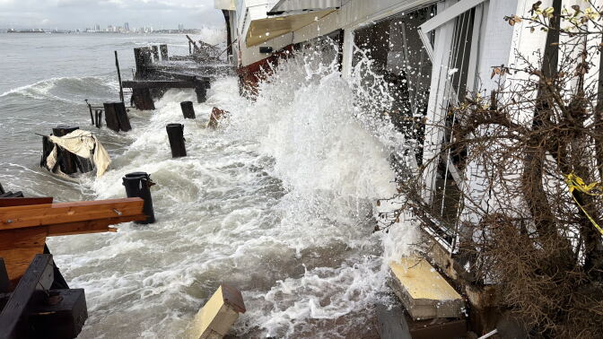 Waves surge against a white waterfront building, splashing up its wall and surrounding broken wooden decking and posts.