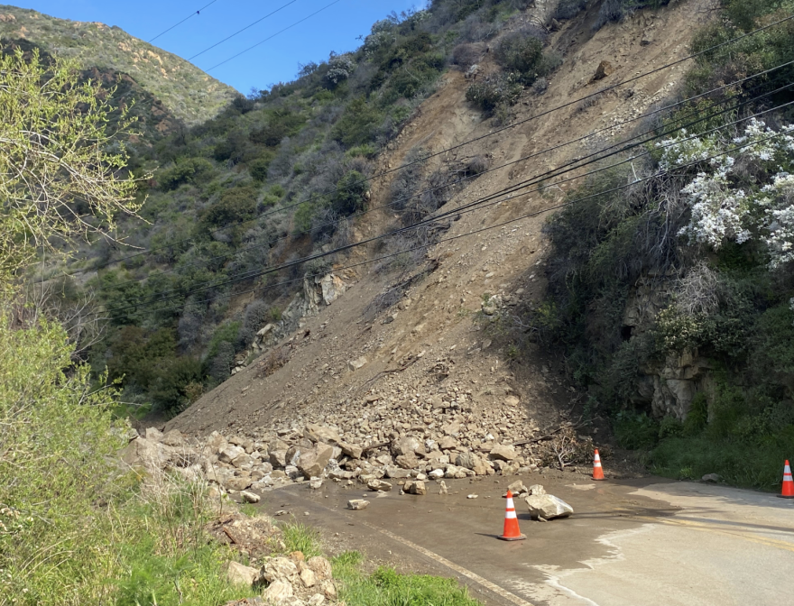 A massive pile of dirt and rocks sits on the roadway having slide from the hillside above. Three orange traffic cones can be seen on the road. Power lines pass through above the roadway.