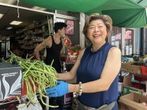 An Asian woman stands at the entrance of a small market under a green tarp, smiling and holding a bunch of long green beans. Behind her, a younger Asian man stands amid stacked produce boxes, and shelves of groceries line the back.
