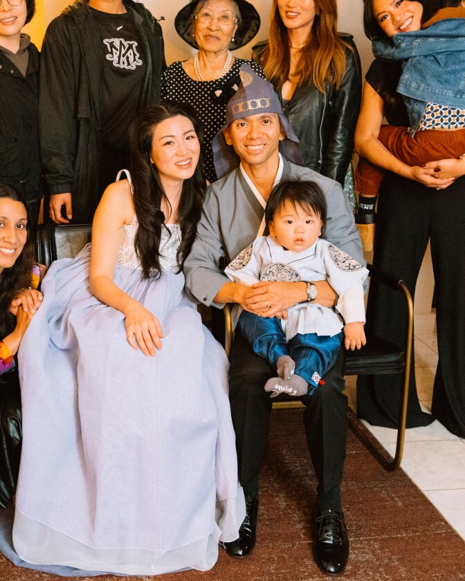 A group photo of nine Asian American male and female adults smiling. A man seated in the chair holds a 1-year-old baby. 