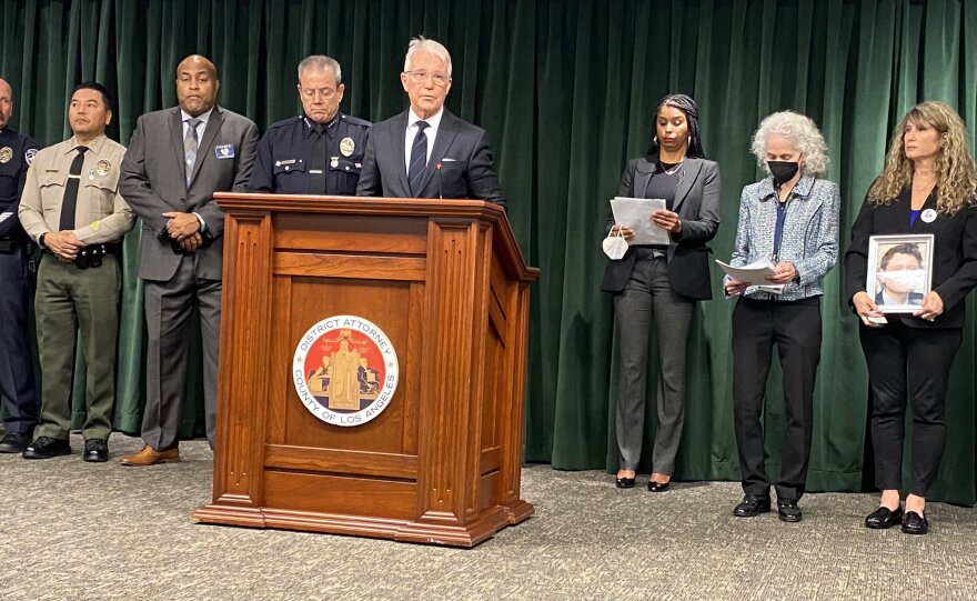 LA County District Attorney George Gascón stands at a wood podium addressing a press conference. He wears a suit and tie and is flanked by other county officials, including Dr. Barbara Ferrer, director of the LA County Dept. of Public Health.