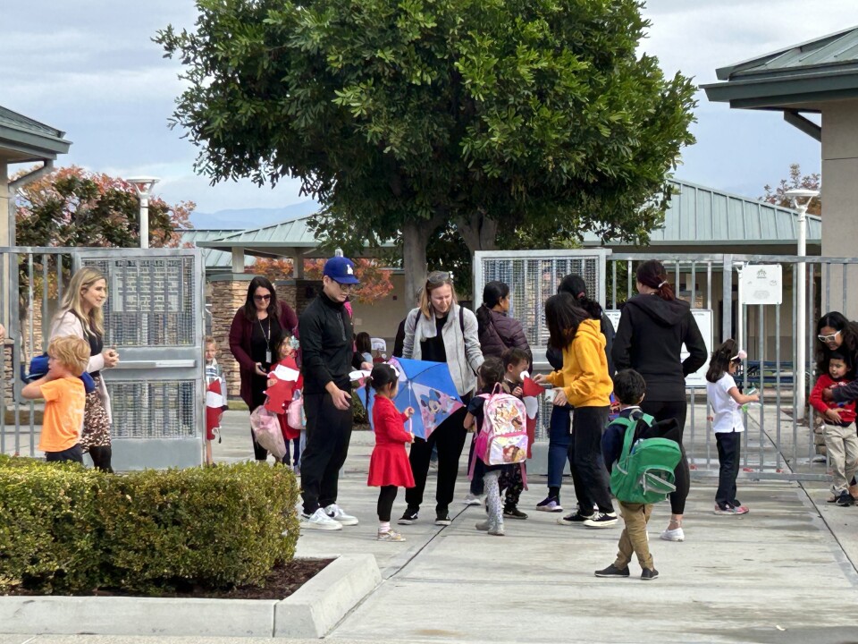 About 18 adults and small children stand near or walk out of a gate in front of a school. Some of the kids are holding cut-out Santas. 