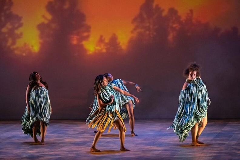 Four barefoot dancers in striped costumes perform on stage with sunset backdrop and warm lighting.