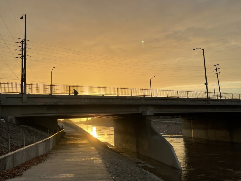 A cyclist cross a bridge at sunset. Beneath him, a shallow river runs through.