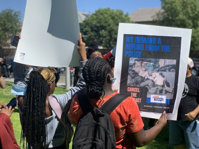 We see two Black students from behind during a protest outside of Lancaster High School in September 2021. The student on the right is wearing their hair in corn rows, has an orange shirt and a black daypack on their shoulders. The student is holding a poster with a white border and a black background on the inside. In blue capital letters at the top left it says "we demand a refund from the police." There is a color photo on the poster of six law enforcement officers pinning someone next to a car. At the bottome left of the poster in orange capital letters it says "cancel the contract." To the right of those words there is a logo that says "cancel the contract." Below the logo in small capital letters are the words "Samuel Chavez Reyes." To the left in the picture is a Black student holding a poster over her head -- we only see the back of the poster, which is all white. The student is wearing a gold scarf on her head, and she has long black hair hanging in braids down much of her back. Sheis wearing a light blue shirt and the light purple strap of a backpack is visible on her right shoulder.