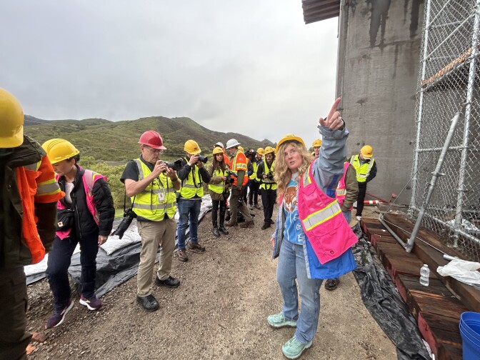 At least eight people, all wearing hard hats and neon high-visibility vests, are throwing handfuls of dirt on the ground in front of them with big smiles on their faces.
