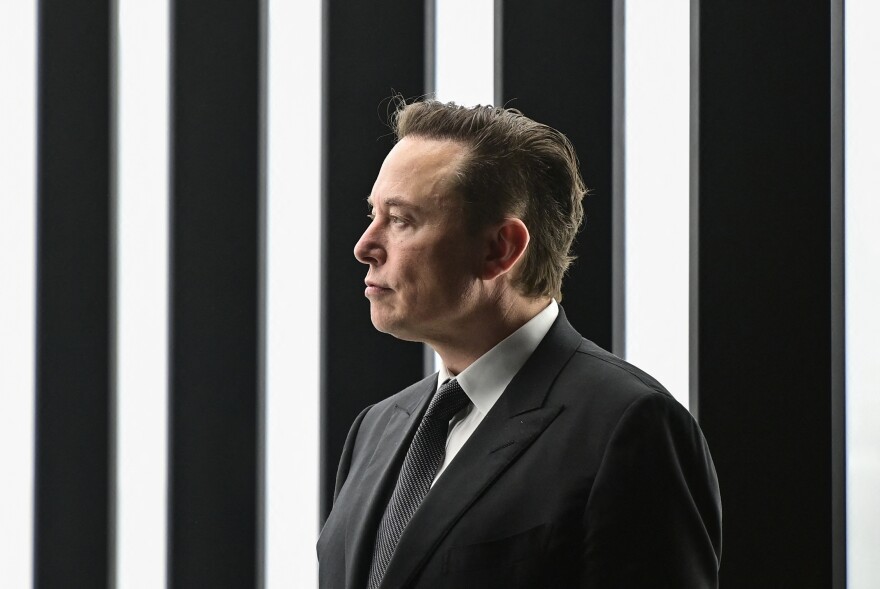 Elon Musk, a 40-something white man, in a dark suit and tie, stands in front of a black-and-white striped background.