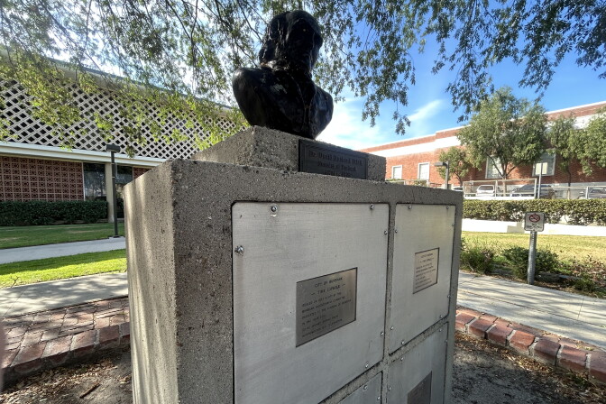 A cement monument with four square compartments topped by a black bust of a man. The monument is outside on a sunny day between two buildings and patches of grass.