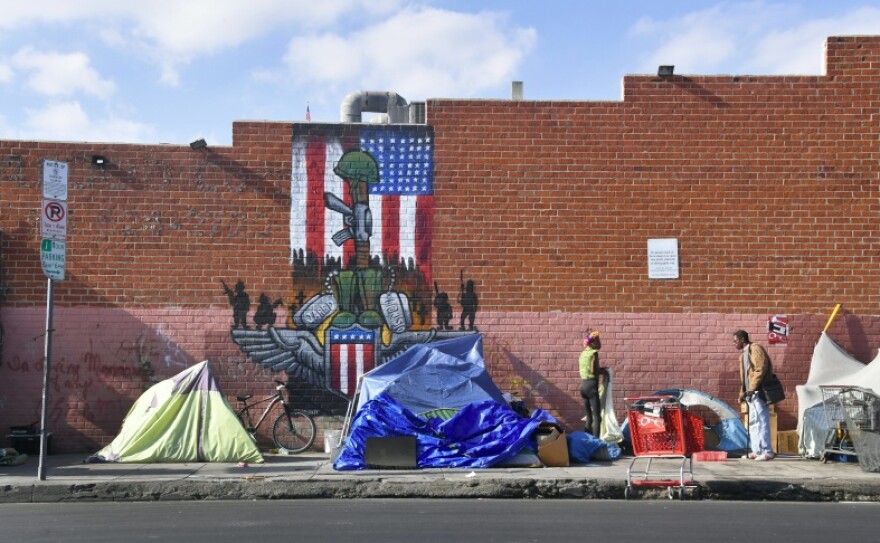 A wider shot reveals a series of tents along the street in front of a brick wall with a mural of the American flag, turned horizontally and also showing a rifle with a helmet perched on its barrel. Two people can be seen around the tents, which have various colors.