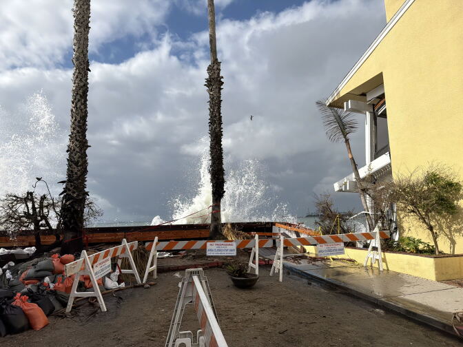 Large waves crash over a damaged wooden seawall behind orange-and-white barricades with “No Trespassing” signs, next to a yellow building.