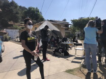 A woman stands wearing all black with a black t-shirt reading "BLM" in yellow. People stand and sit behind her in a tent and on chairs on a residential street. 