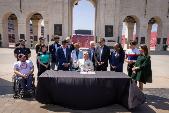 A group of people, including several in USA Olympic and Paralympic team apparel, gather around a table at the entrance arch of the Los Angeles Memorial Coliseum for a document-signing ceremony. A seated official signs papers at the black-draped table while others look on and smile, with the stadium's stands and bronze plaques visible in the background.