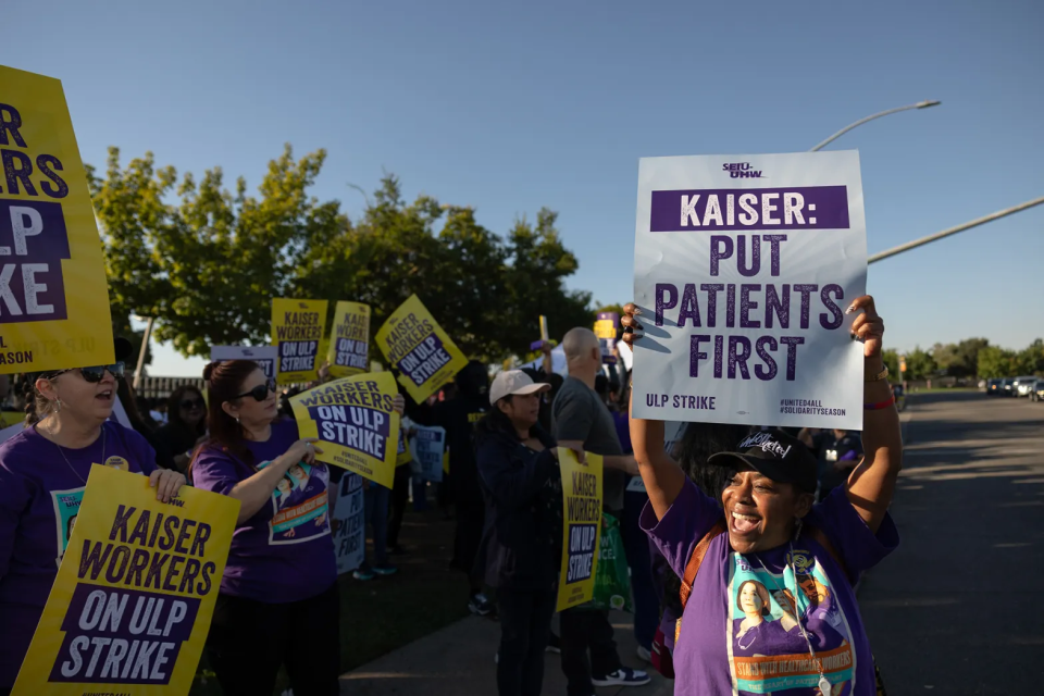 A woman with brown skin, wearing a baseball cap and a purple t-shirt, holds a poster over her head that reads Kaiser: Patients First. Beside her are dozens of other protesters, most holding posters that read Kaiser Workers On ULP Strike