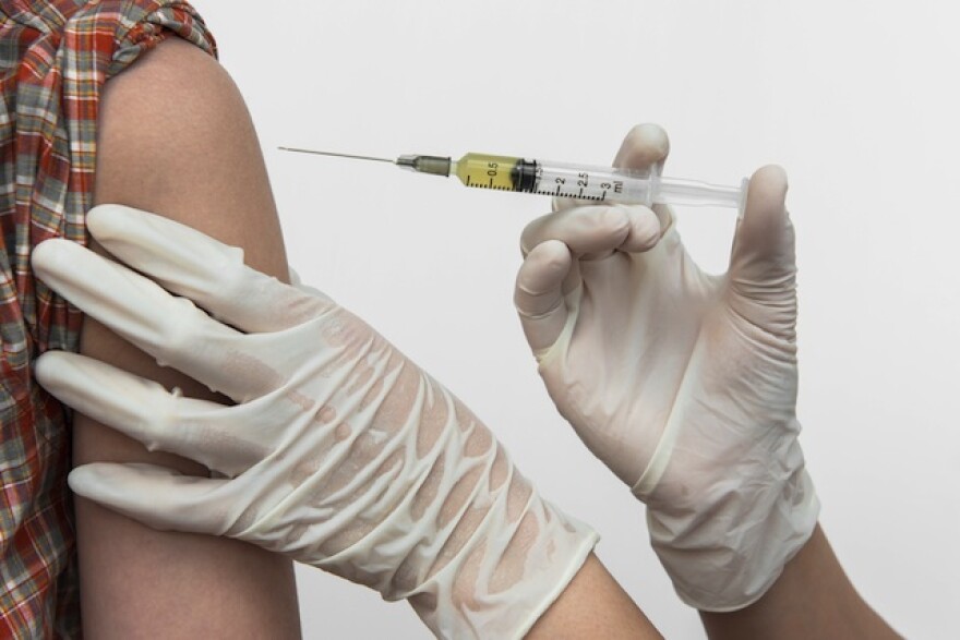 A person holds a syringe filled with a vaccination. Only the hands of the person holding the syringe is visible, they are wearing white medical gloves and slightly pushing down with their right hand. The syringe hovers above an arm. The person receiving the vaccine is wearing a red, grey and white plaid shirt.
