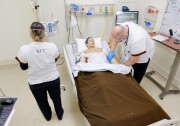 A man in a white uniform, bends over a hospital bed and examines a mannequin's arm. A woman stands next to the bed with her back to the camera. Her white uniform has the letters "RCC" on it.