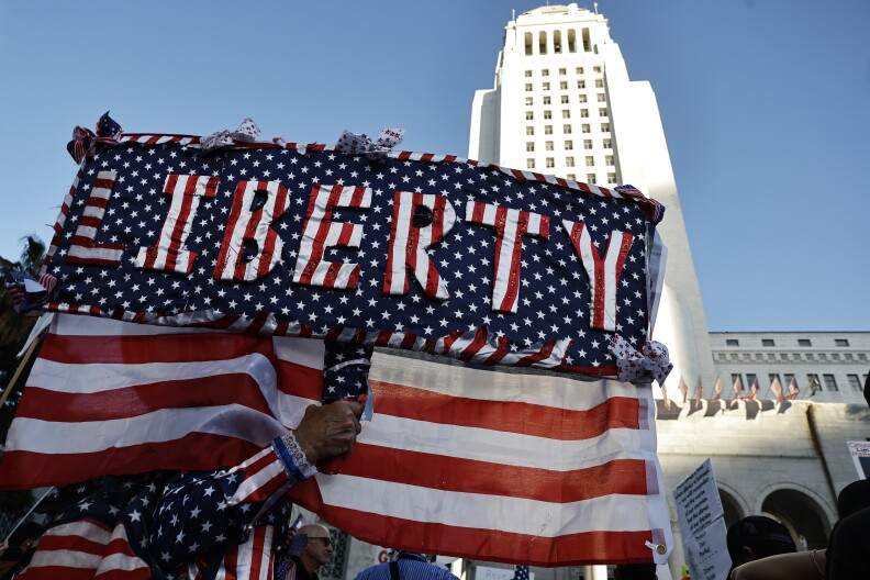 Red white and blue fabric reads: Liberty in a sign carried with a U.S. flag. L.A.'s city hall is visible in the background
