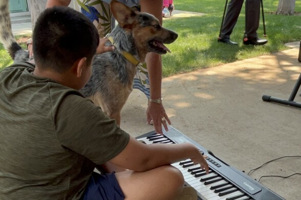 A boy wearing a dark green t-shirt and blue gym shorts is sitting on the cement ground with a portable keyboard in front of him. A brown and black cattle dog is standing to his left, with a woman leaning over the pair.