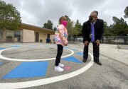 A man in a navy suit and purple tie bends down to chat with a young girl wearing a shiny pink coat. They are on a cement surface outside a one-story school building, and both are wearing masks.