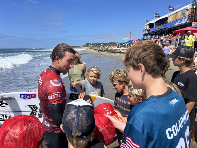 A wetsuit-clad surfer signs hats and a surfboard for a group of children gathered on the beach near a competition venue. One of the kids has a shirt that reads "Colapinto" on the back. 