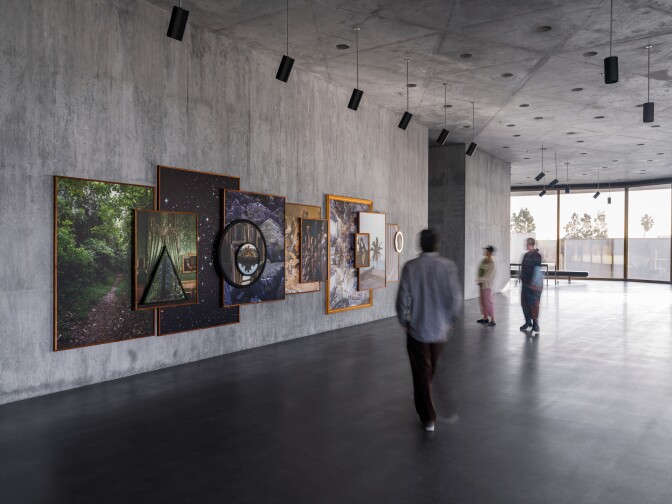 People stand in a large room with grey concrete walls. Art hangs on the walls, and there is a general sense of light from the floor to ceiling windows.