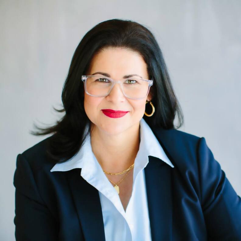 a headshot of a woman with brown hair, glasses, a white blouse and a blue jacket