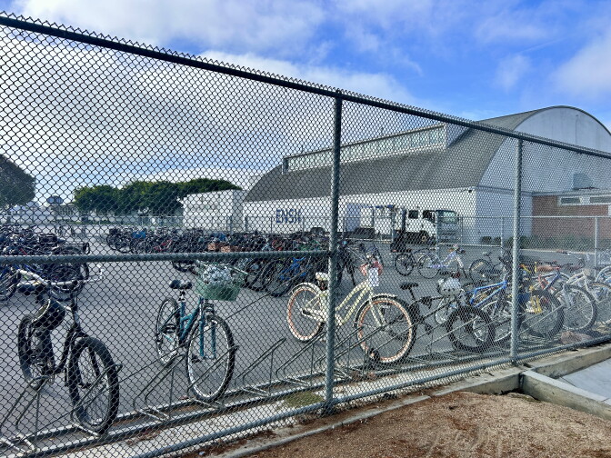 Dozens of bicycles are parked behind a wired fence next to a school gym. 