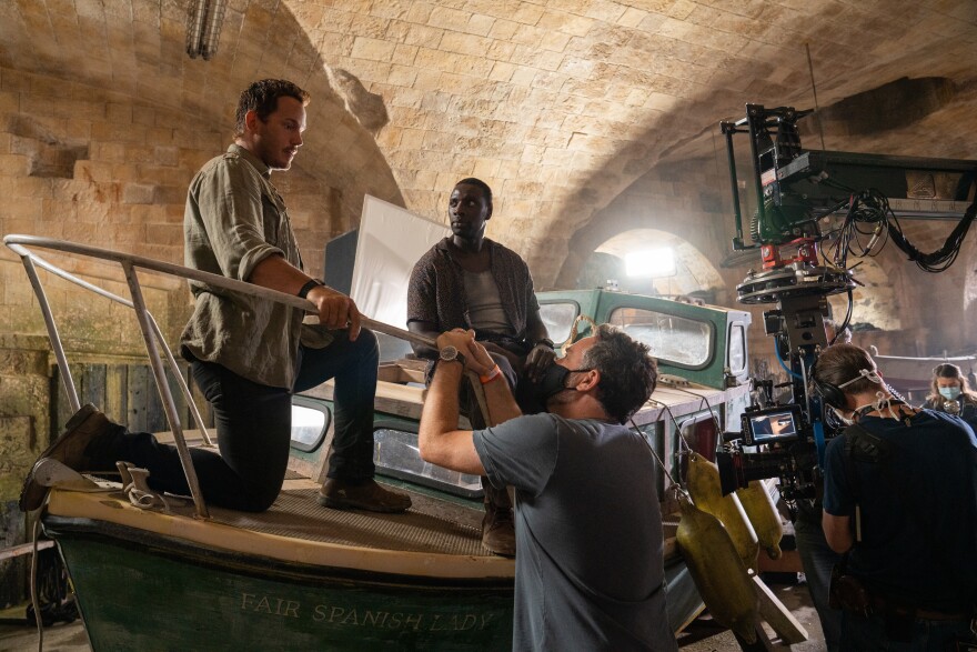 Two unmasked men sit on a boat as they talk to another masked man leaning over the side from the ground. A film camera is at the right.