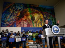 Gavin Newsom stands at a lectern with his left arm raised. In the background is a large-format mural of showing a person holding a diploma, people dancing and basketball game.