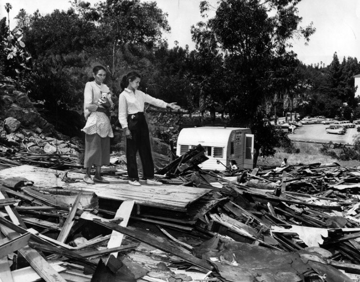 Two people stand on a pile of debris. One is gesturing at the mess.
