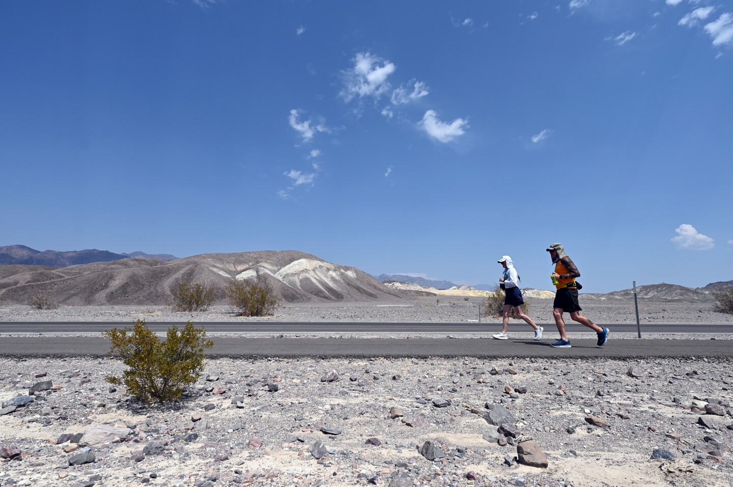 Two runners make there way on roadway in Death Valley, surrounded by desert terrain.