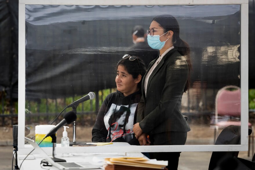 Two women, one sitting and the other standing with a mask on, behind a table with a microhphone.