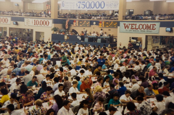 A wide look at a large room that's packed with rows of people sitting at tables playing bingo. There are banners hanging on the walls showing welcome and prize amounts, such as one for $10,000.