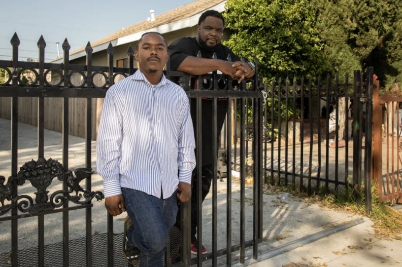 Two men with dark brown skin tone are leaning against a black iron gate. One is wearing a light shirt and jeans, and the other appears to wearing a black t-shirt and pants.