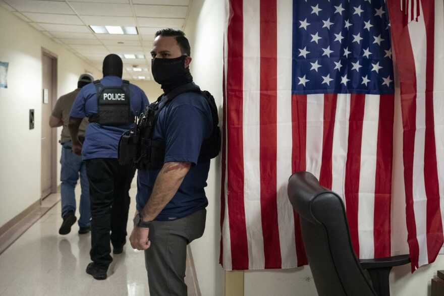 Federal agents stand and walk in a hallway. One agent with medium skin tone, wearing a black mask, stands by a wall where an American flag hangs vertically.