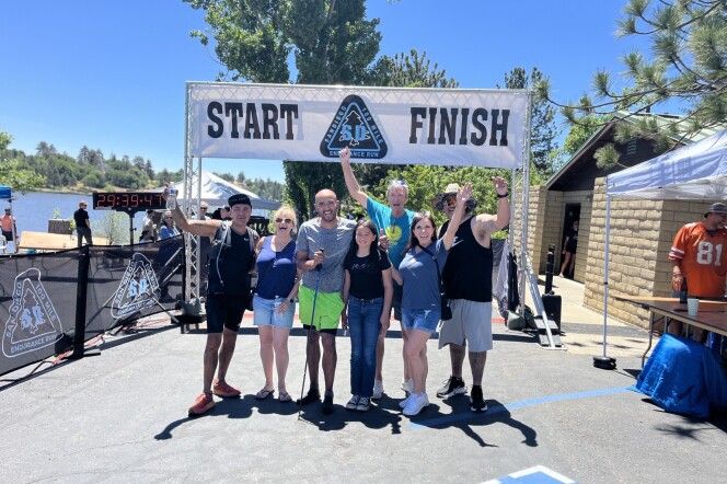 A group of people stand at a finish line with their arms around each other and up in the air. They are smiling.