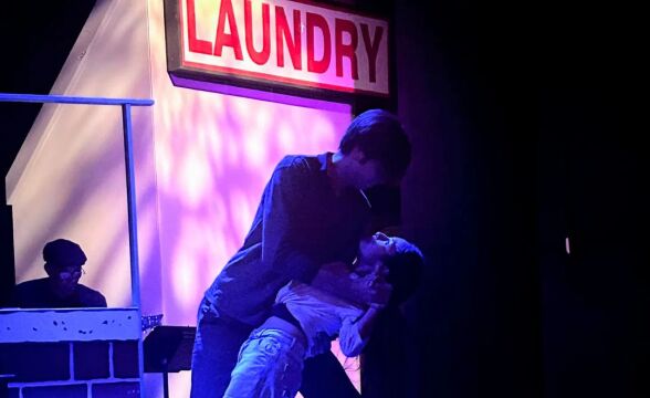 A man dips a woman at the end of a dance, both of them bathed in blue light. Above them is a sign that says "Laundry." 