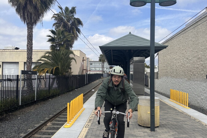 A young man with a white bike helmet and wearing a jacket glasses sits on his bike on a Metro platform